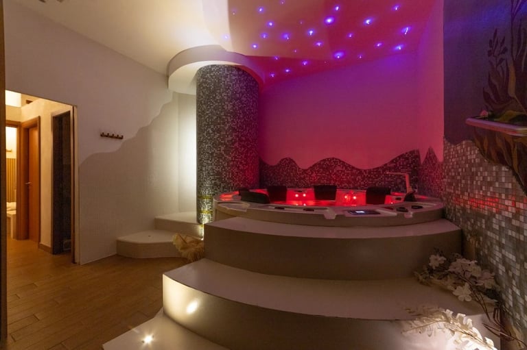 Modern spa room with circular sunken seating area, purple and pink ambient lighting, decorative stones, and a textured cylindrical column in the center